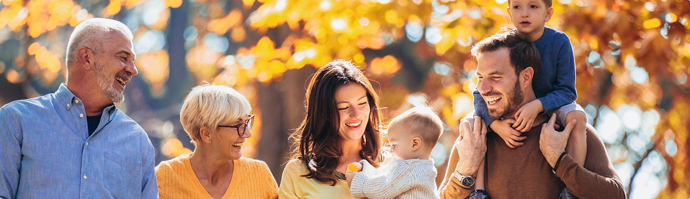 Three generations of a family smiling outdoors with autumn trees in the background
